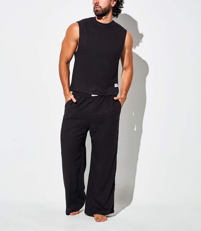 Man wearing a sleeveless black top and loose black pants, standing barefoot.