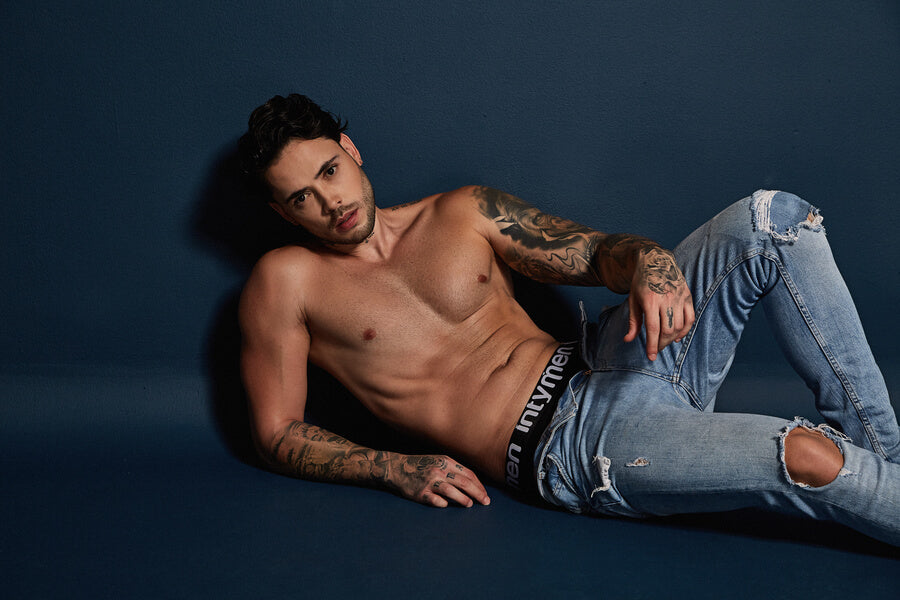 Do mens thongs count to be the perfect sleepwear -  Shirtless, tattooed man in ripped jeans reclining on a blue background