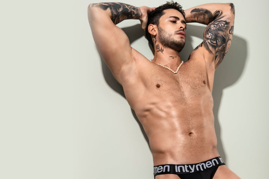 Earn bonus points this summer with Intymen's Men's Bikinis - Shirtless, tattooed man in black underwear posing with hands behind head 