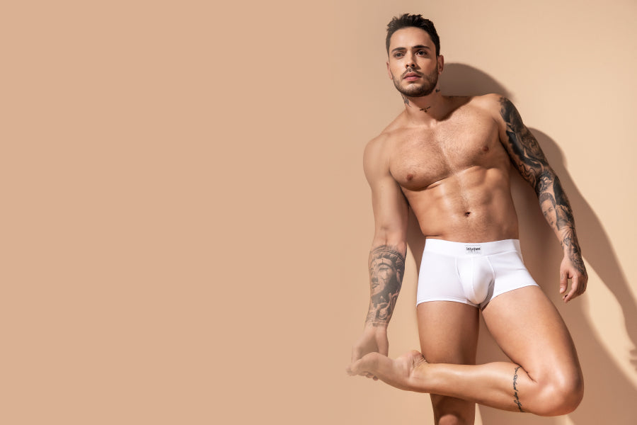 I didn't know men's mesh underwear can be incredible too - Muscular, tattooed man in white boxer briefs posing against a beige wall 