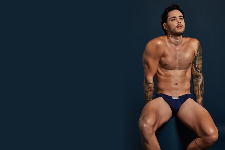 Who doesn’t like getting strapped on? - Man sitting and posing in navy underwear against a dark blue background.